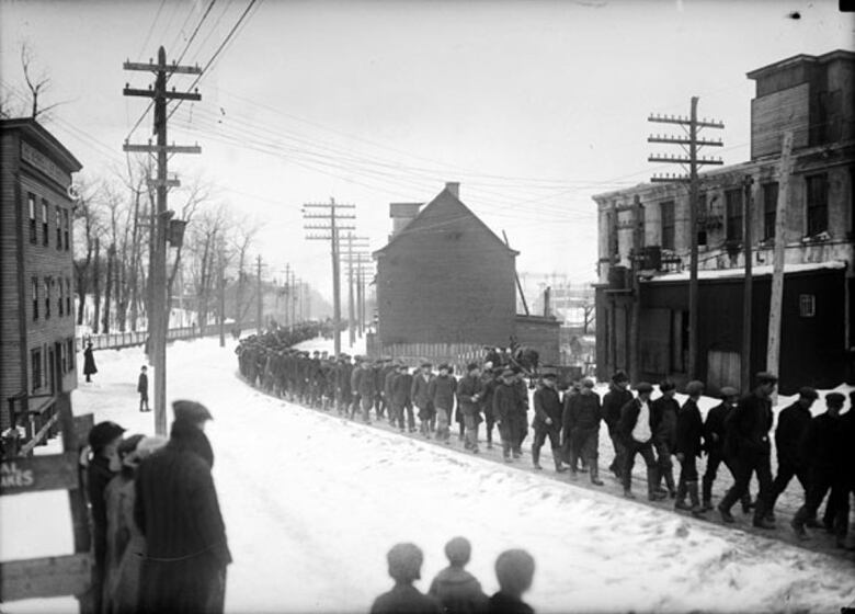 An old black and white photograph of a long line of men in dark clothes marching down a town road.