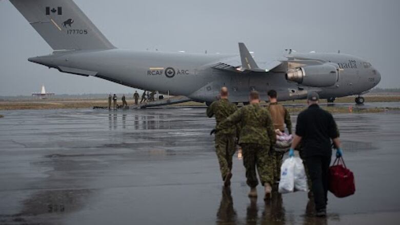 Four men, two of them in green army uniform, carry a stretcher on the tarmac towards a large, grey military plane.