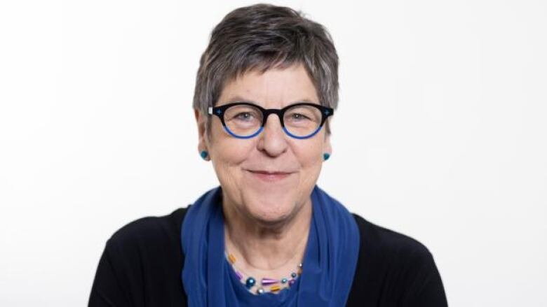 An older woman with short grey hair wearing blue rimmed glasses and blue earrings. She is wearing a black top with a blue scarf around her neck