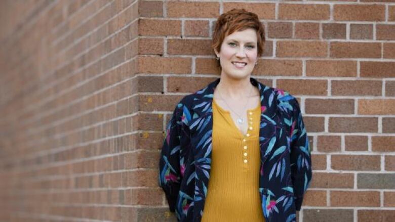 A photo of the author who is a woman with short red hair. She is wearing a mustard yellow shirt with a blue cardigan that has tropical leaves on it. She is leaning against a brick wall and smiling at the camera. 