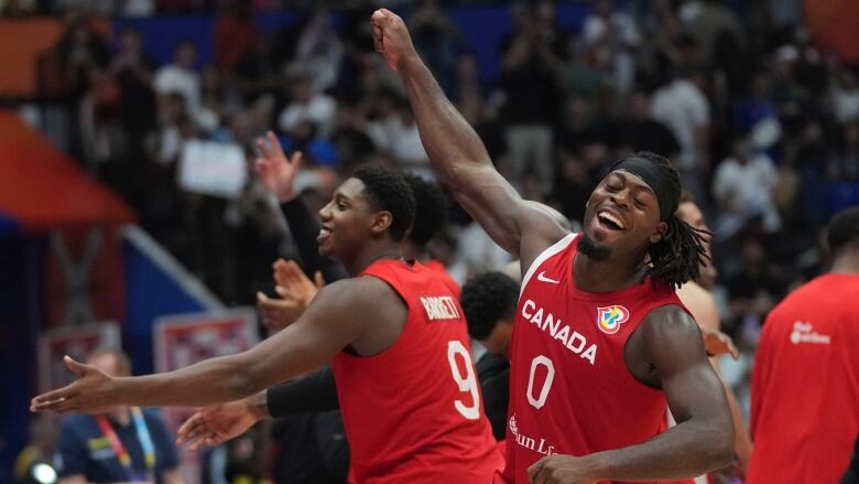 Two men's basketball players, wearing red jerseys with white lettering, throw their arms in the air in celebration.