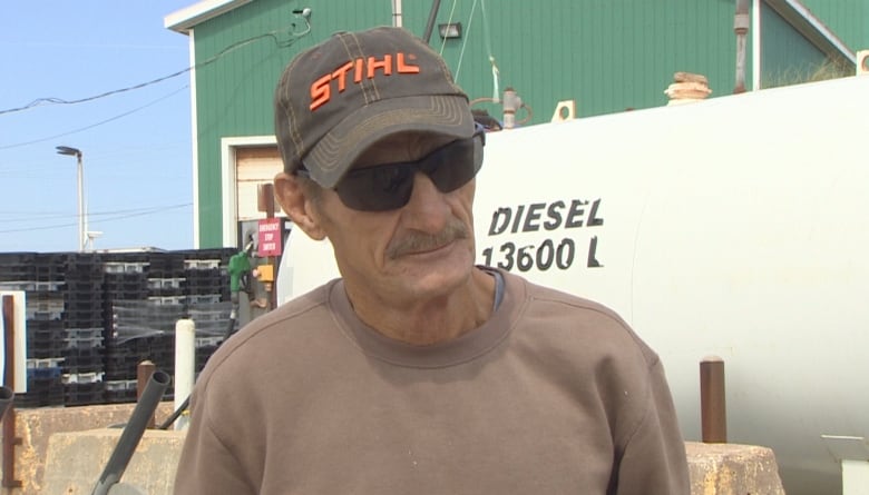 Older man in sunglasses and a ball cap stands in front of a diesel tank. 