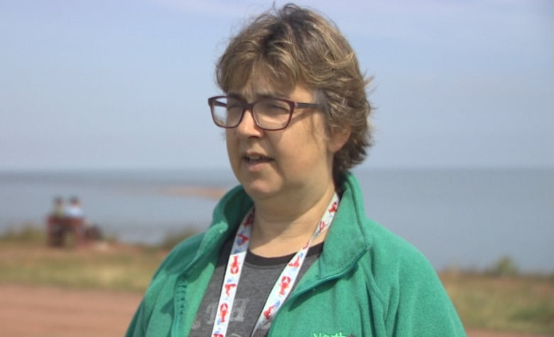 Woman with short hair and glasses stands on a cliff overlooking the ocean. 