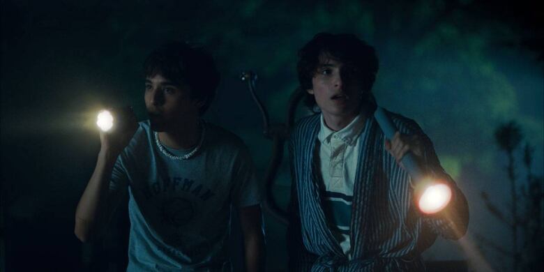 Night time scene. Two white young men with floppy dark hair, Finn Wolfhard and Billy Bryk, hold flashlights and appear to walk through a dark wood.