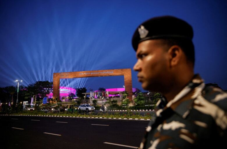 A security officer with a black beret stands guard in front of an archway in front of a stadium lit up with pink lights at night.