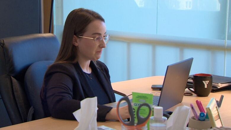 A woman with long brown hair sits at her desk working on a laptop