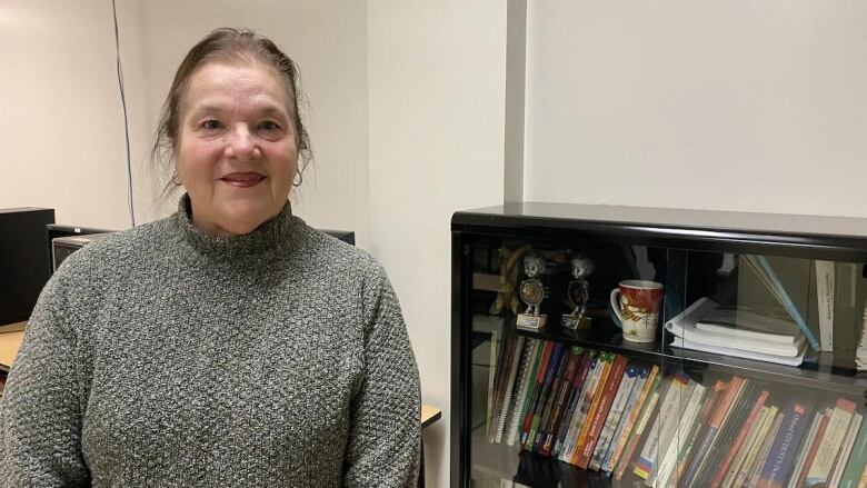 A woman is seated indoors to the left of a black book shelf with glass doors. She has her hair tied back and is wearing a grey textured turtle neck sweater.