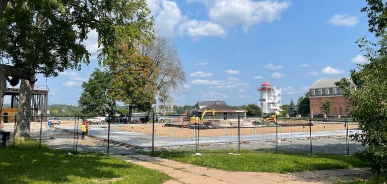 A panoramic shot of Officers' Square from the permanent stage under construction at left across the square where a rectangular concrete track has been laid. The lighthouse can be seen in the background. Some leafy tree branches frame the foreground.