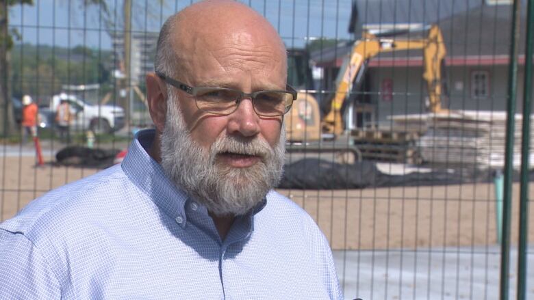 A head and shoulders shot of a man with a white beard wearing a light blue button-down collared shirt who is standing in front of some heavy equipment at a construction site.