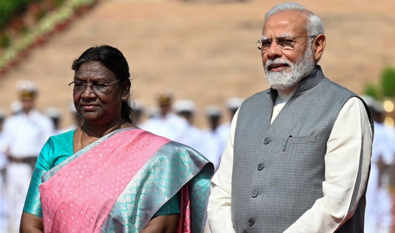 Three people, two men in suits and a woman in a pink and turquoise sari, stand on a stage outdoors as they pose for a picture.