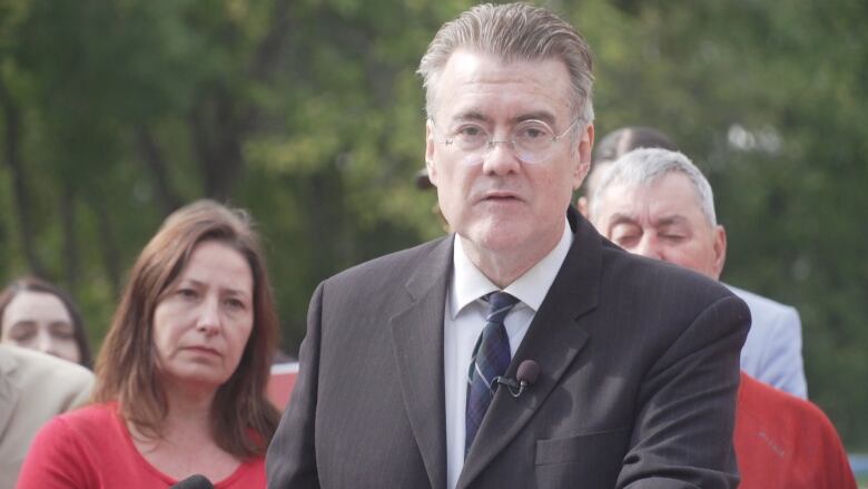 A man in a suit and tie speaks at a microphone, while flanked by other people.