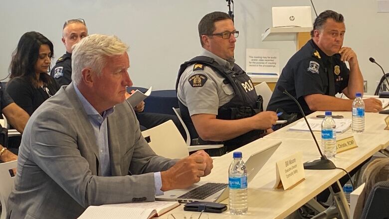 Three men sit behind a table with microphones on top of it, two of them are wearing police uniforms. 