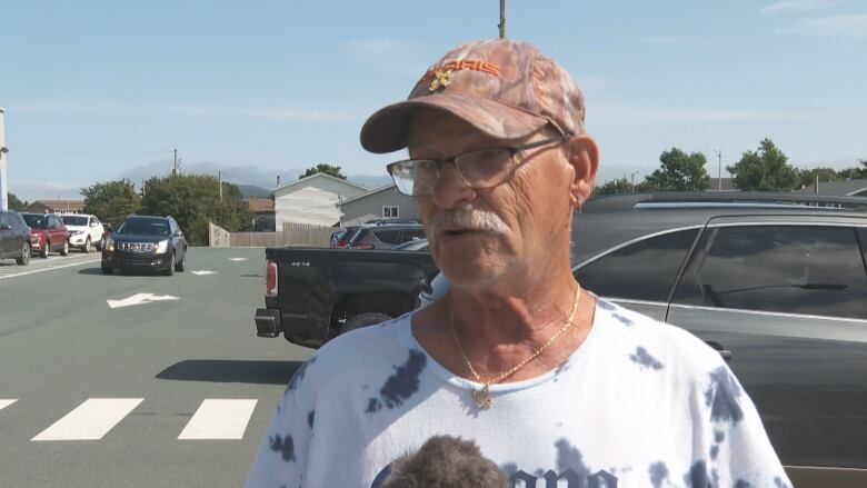 middle aged man wearing a hat and glasses stands in a supermarket parking lot.