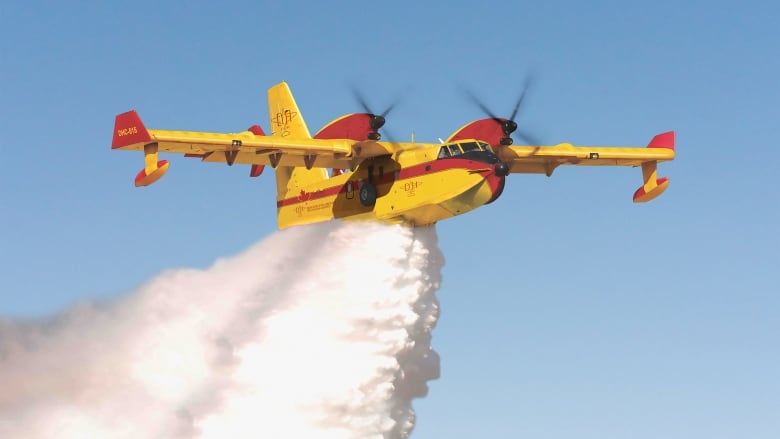 A yellow plane dropping a plume of water flies against a blue sky