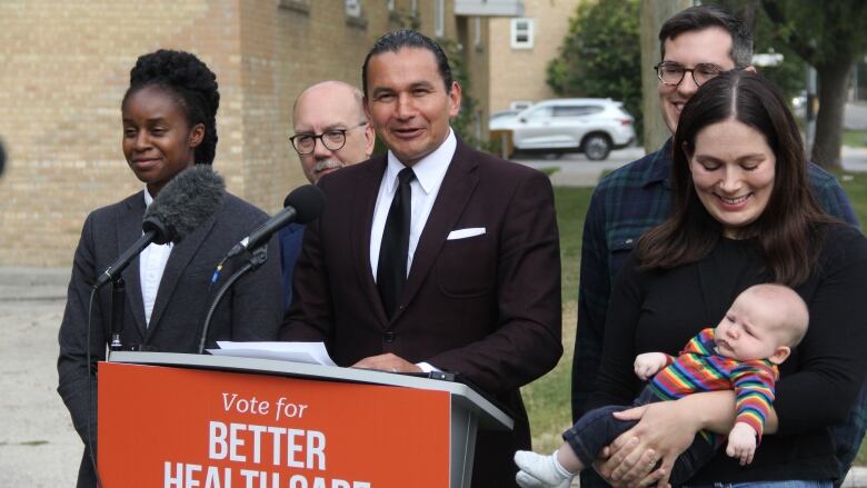 A man in a suit and tie stands behind a podium, while other people stand around and behind him.