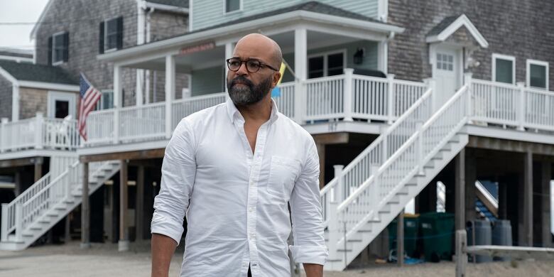 Actor Jeffrey Wright wearing a white-collared shirt stands in front of a beach house with a wrap-around porch and an American flag.