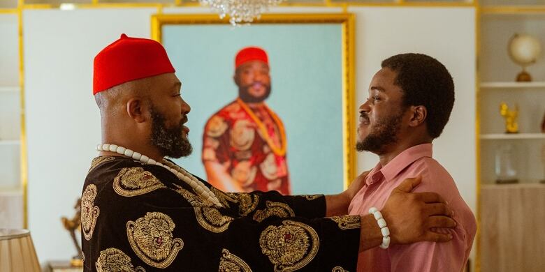 Two Black men stand facing each other. One is wearing a red hat, white-beaded necklace and a black and gold robe. The younger Black man is wearing a salmon collared shirt.