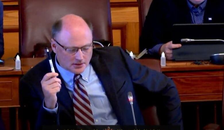 A man wearing glasses and a suit, speaking into a microphone while holding a pen in the air.