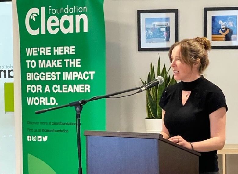 A woman stands at a podium giving a speech. She's speaking into a microphone. A green poster in the background reads: 