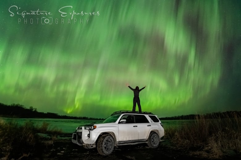 Woman stands atop a white sport utility vehicle facing the aurora borealis.