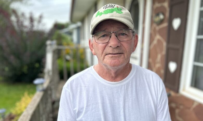 A man in a white T-shirt with ballcap and glasses stands on his porch