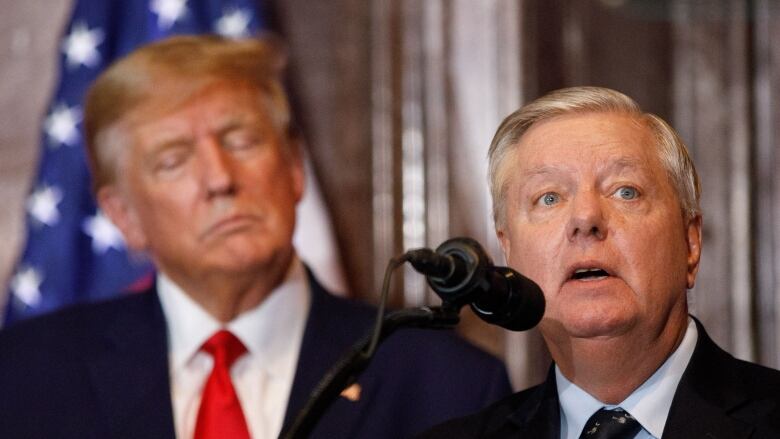 A man in a suit and tie speaks into a microphone as another looks on.