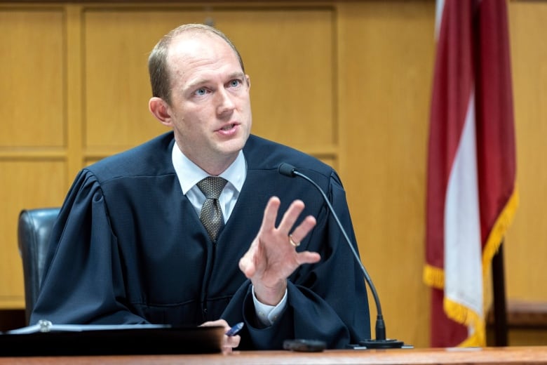 A judge wearing a robe over a collared shirt and tie is shown speaking into a microphone at the bench in a courtroom.