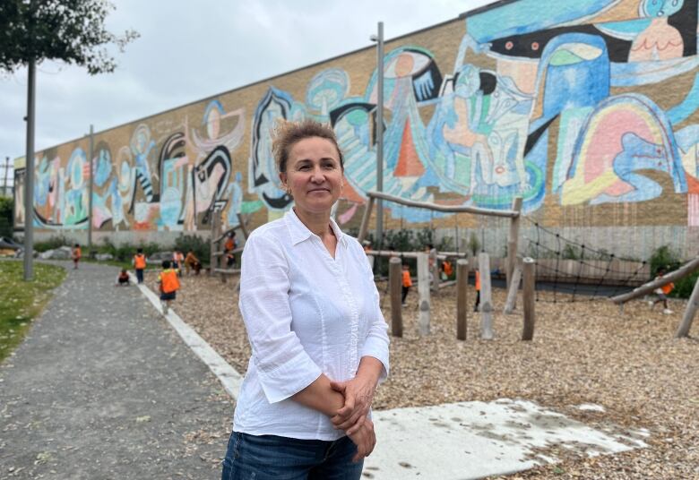 A woman stands in front of a playground backed by a mural where young children are playing.