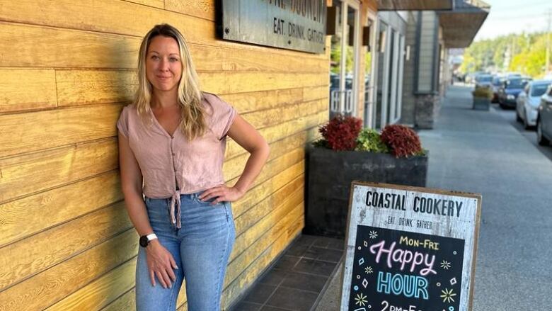 A woman smiles and leans against a wood building, with a sign beside her saying it is a restaurant with a happy hour.