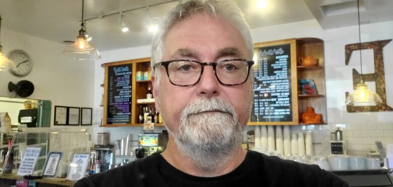 A man with glasses and a beard is in a cafe with a menu behind him and a serious or concerned look