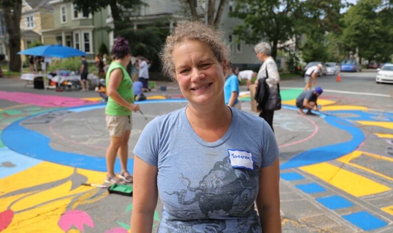 A woman wearing a name-tag that says Susanna smiles at the camera. People can be seen painting a bright mural on the street behind her.