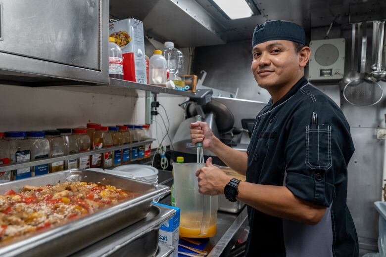 A man wearing an apron and cook's cap smiles while standing in a kitchen area and holding up a jug of sauce that he is stirring. 