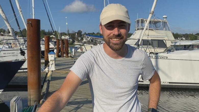 A man wearing a tshirt and a ball cap stands on a dock with boats behind him.