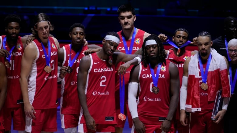 Several men's basketball players, all wearing red jerseys with white lettering, pose with their medals.