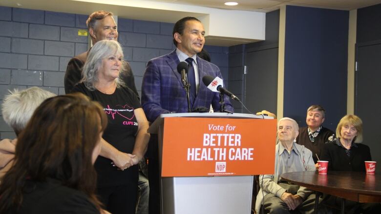 A man speaks at a podium, surrounded by a few people standing beside him and other people seated around a few tables