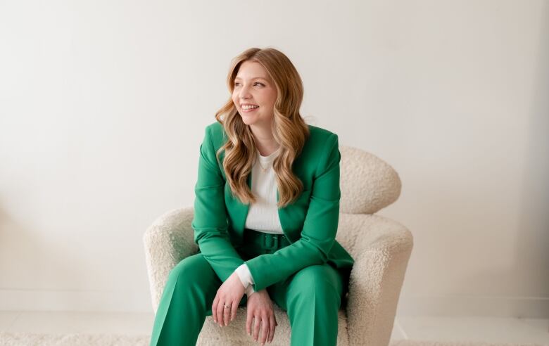 A woman with long, lightly curled hair, wearing a bright green suit, smiles as she sits in a white chair, with her wrists crossed in front of her.