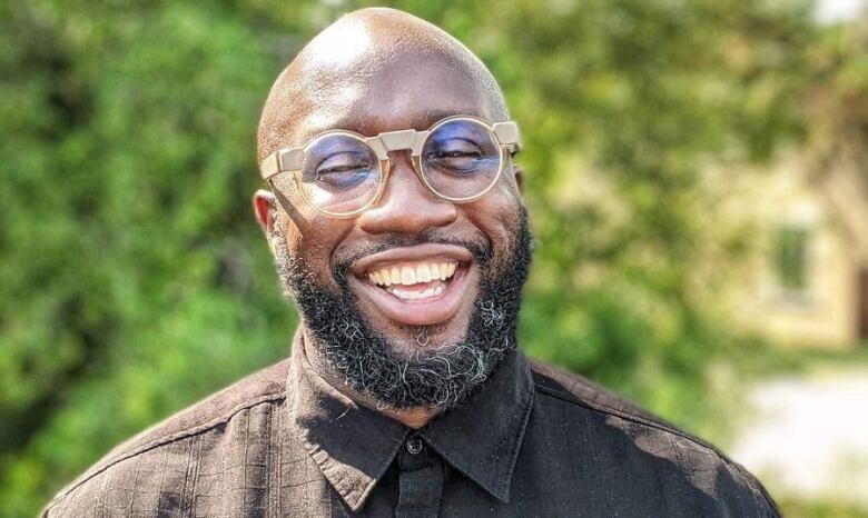 A man with a bald head and black and grey beard, wearing gold glasses and a brown button-up shirt, smiles broadly while standing outside.