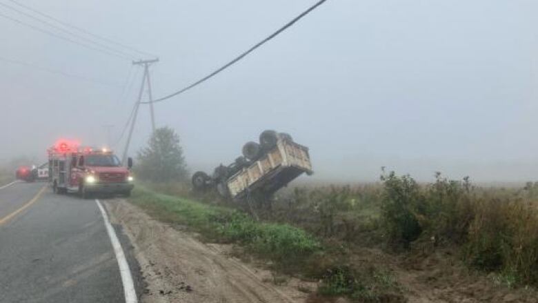 A dump truck or something similar lies on its roof off the side of the road on a foggy day.