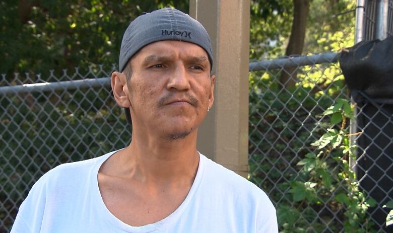 A man standing outside in front of a fence, wearing a white t-shirt and grey backwards cap.