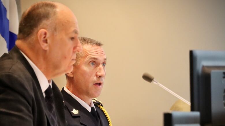 A man in a navy blue uniform with gold braid and a gold maple leaf speaks into a microphone.