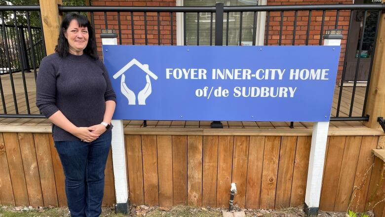 A woman in a black sweater standing next to a blue sign that says Inner City Home of Sudbury.