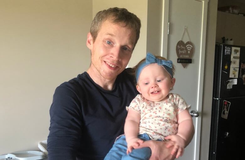 A man smiles, holding a baby who wears a blue bow in her hair.