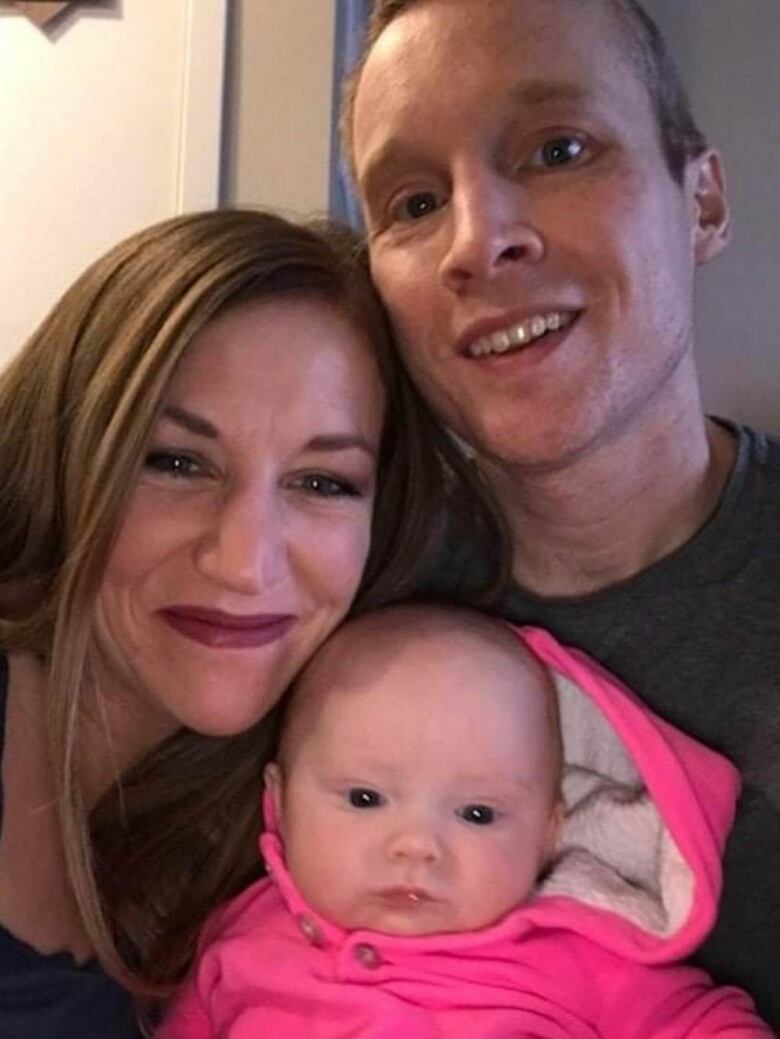 A young family, including a mom, dad and baby take a selfie. The parents are smiling. The baby wears a bright pink onesie.