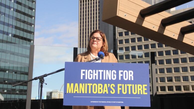 A woman speaks behind a podium, on the second-floor of an outdoor terrace as downtown skyscrapers are seen behind. 