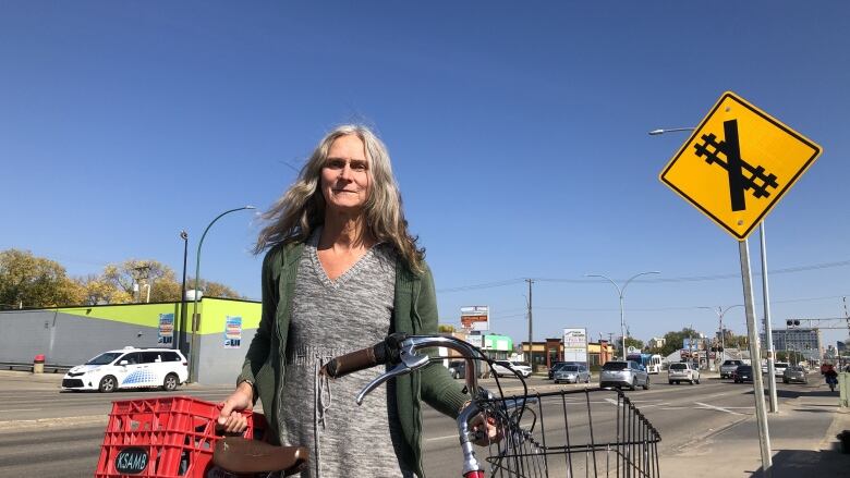 A woman can be seen wearing a grey dress and a green sweater as she stands next to her bicycle which is red and has baskets both in the front and back. 