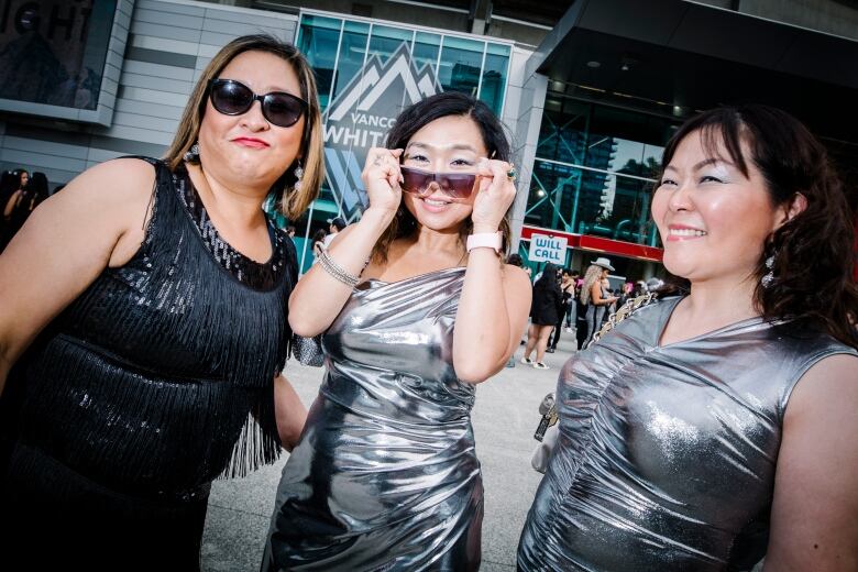 Three older East Asian woman, mostly clad in silver, pose in front of a stadium.