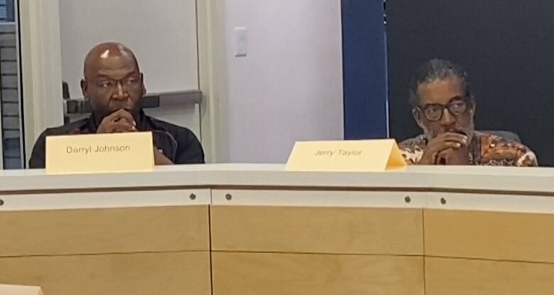 Two Black men sit beside one another at a long white table with microphones