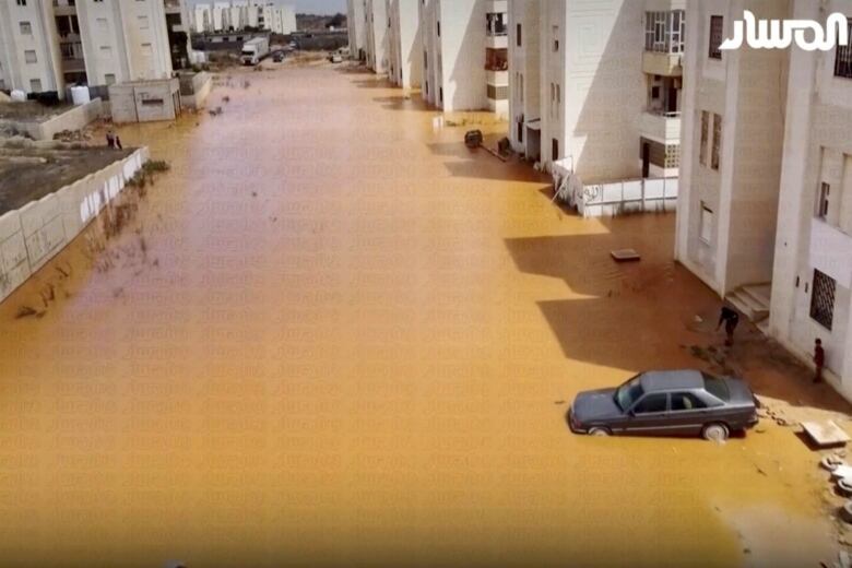 Muddy brown water is shown filling up a street, partially submerging a vehicle and climbing up the sides of buildings.