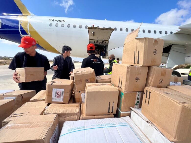 Several people are shown, two in baseball caps, with a pile of boxes on the tarmac near an airplane.