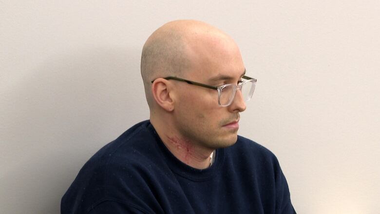 A bald man wearing glasses sits in a prisoners box inside a court room. 